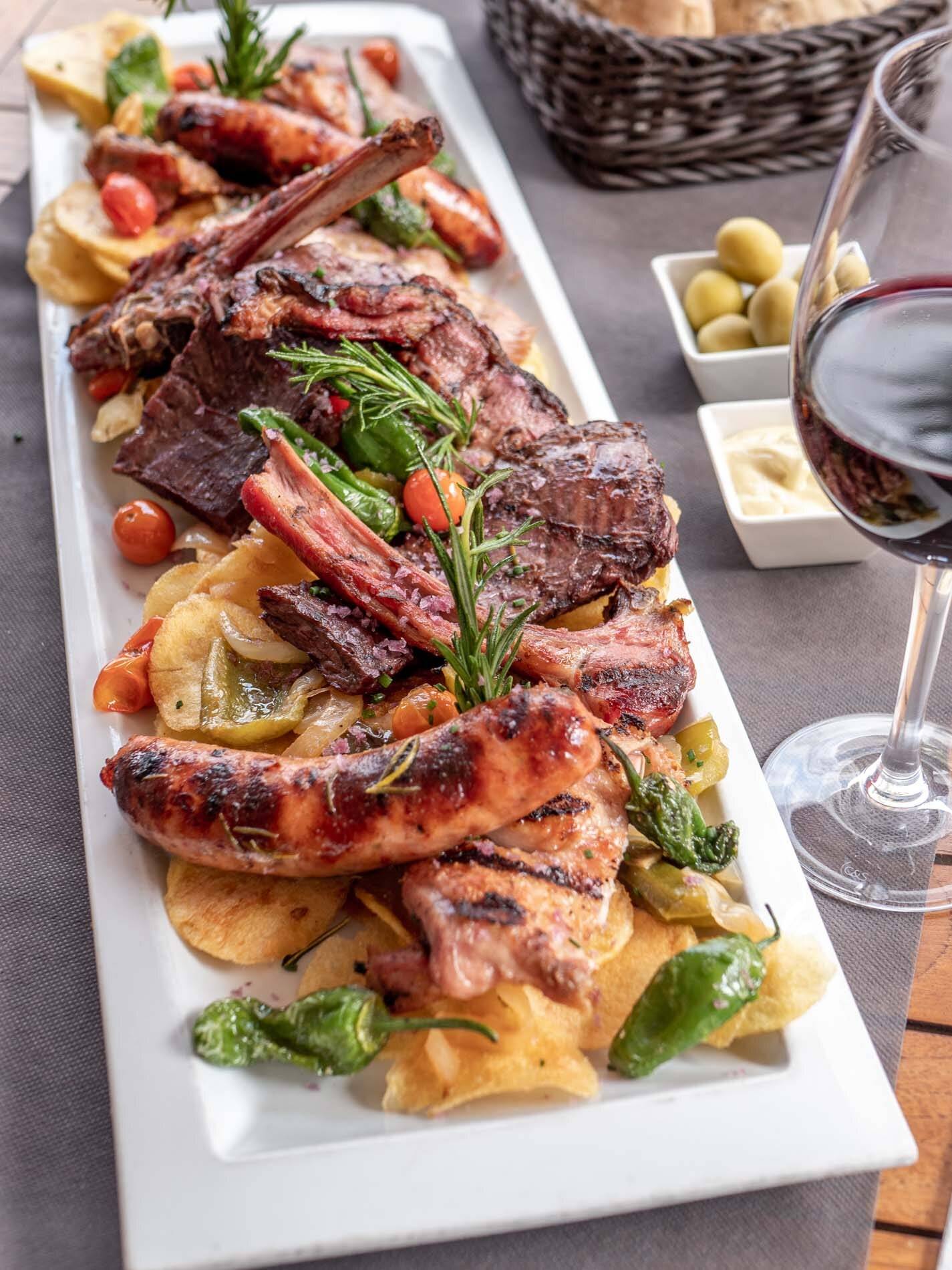 Parrillada de carnes a la brasa con chuletas, costillas y salchichas sobre patatas, con pimientos de Padrón y una copa de vino tinto